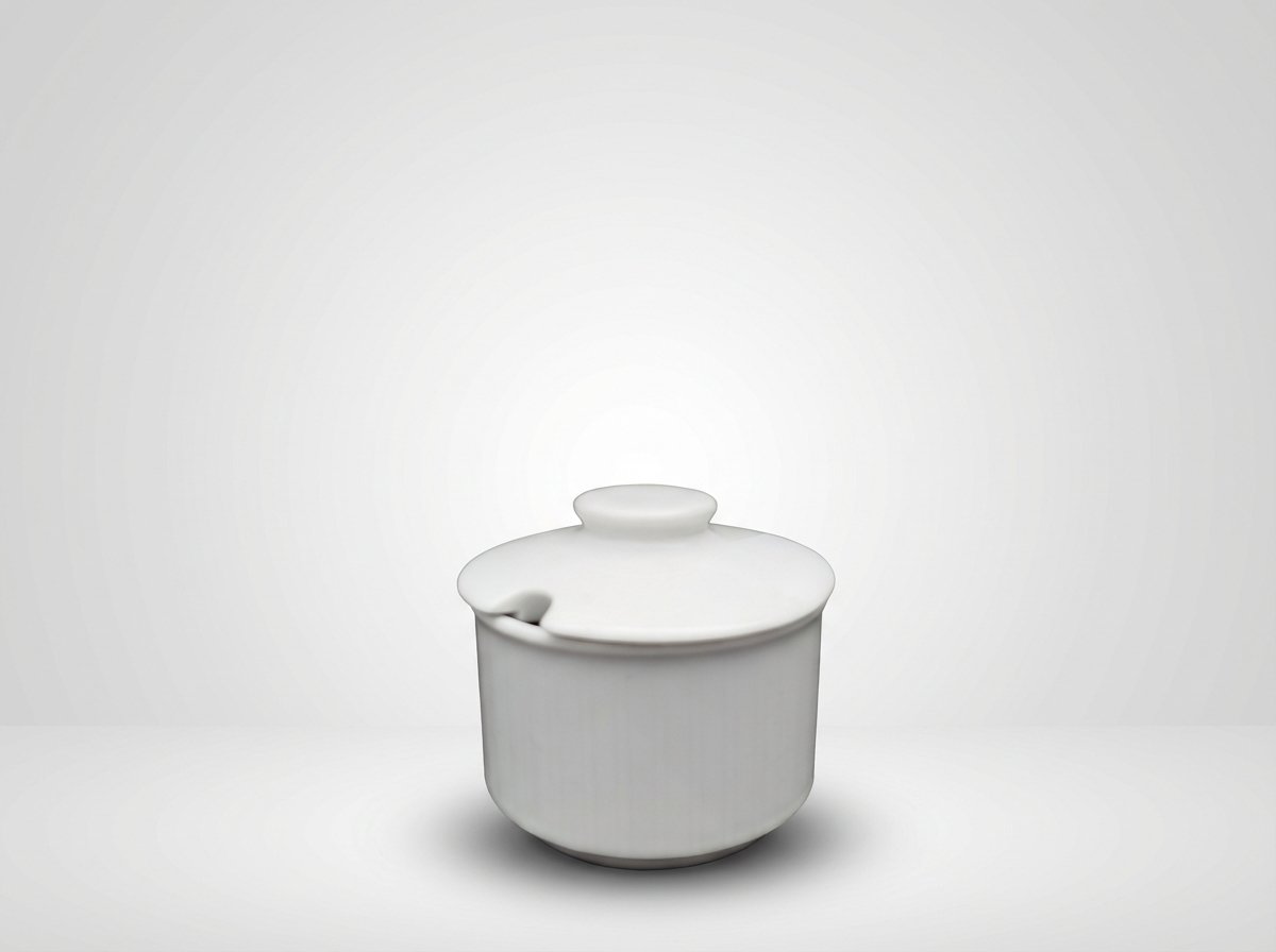 Balta keraminė indelė su dangteliu ir specialia išpjova šaukšteliui, stovinti ant šviesaus pagrindo.