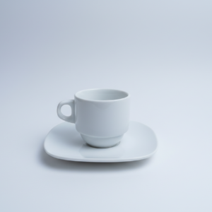 Minimalistinio dizaino balta puodelio ir lėkštutės komplektas, stovintis ant balto paviršiaus ir traukiantis akį savo paprastumu.
