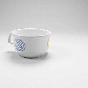 Baltas puodelis 235 ml su mėlyna ir geltona spiralėmis, stovintis ant šviesiai pilko paviršiaus.